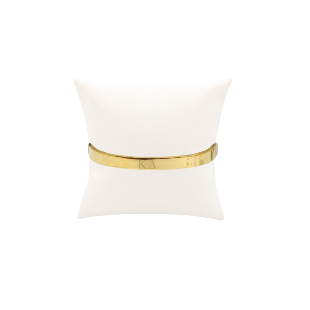 Kappa Delta gold bangle bracelet with greek letters on white pillow front view.
