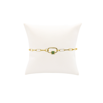 A gold paperclip chain charm bracelet with green cubic zirconia on a white display stand.
