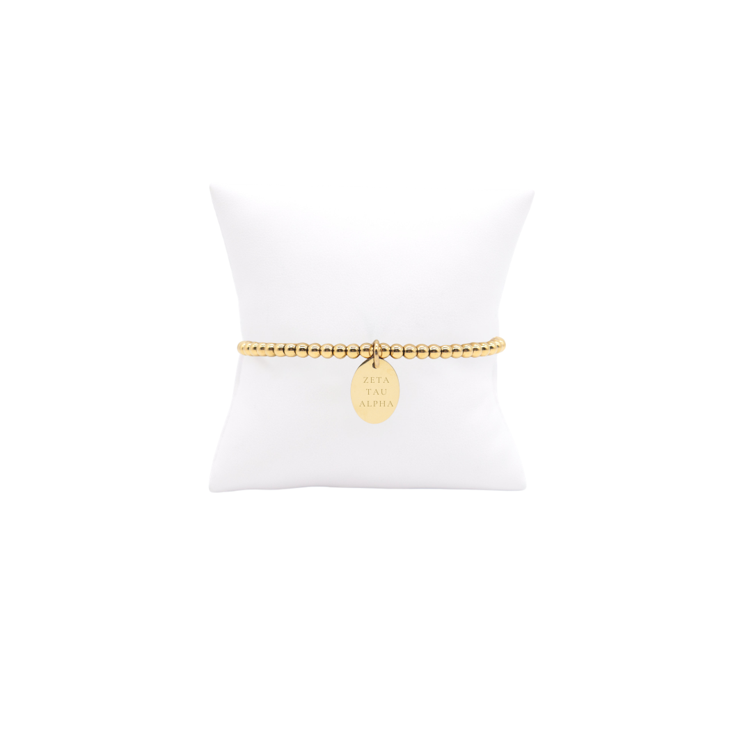 Zeta Tau Alpha gold beaded bracelet with small oval charm with full sorority name displayed on white pillow front view