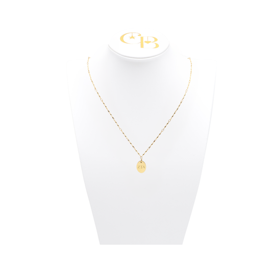 Zeta Tau Alpha gold twist necklace with small oval charm with horizontal greek letters on white pillow front view.