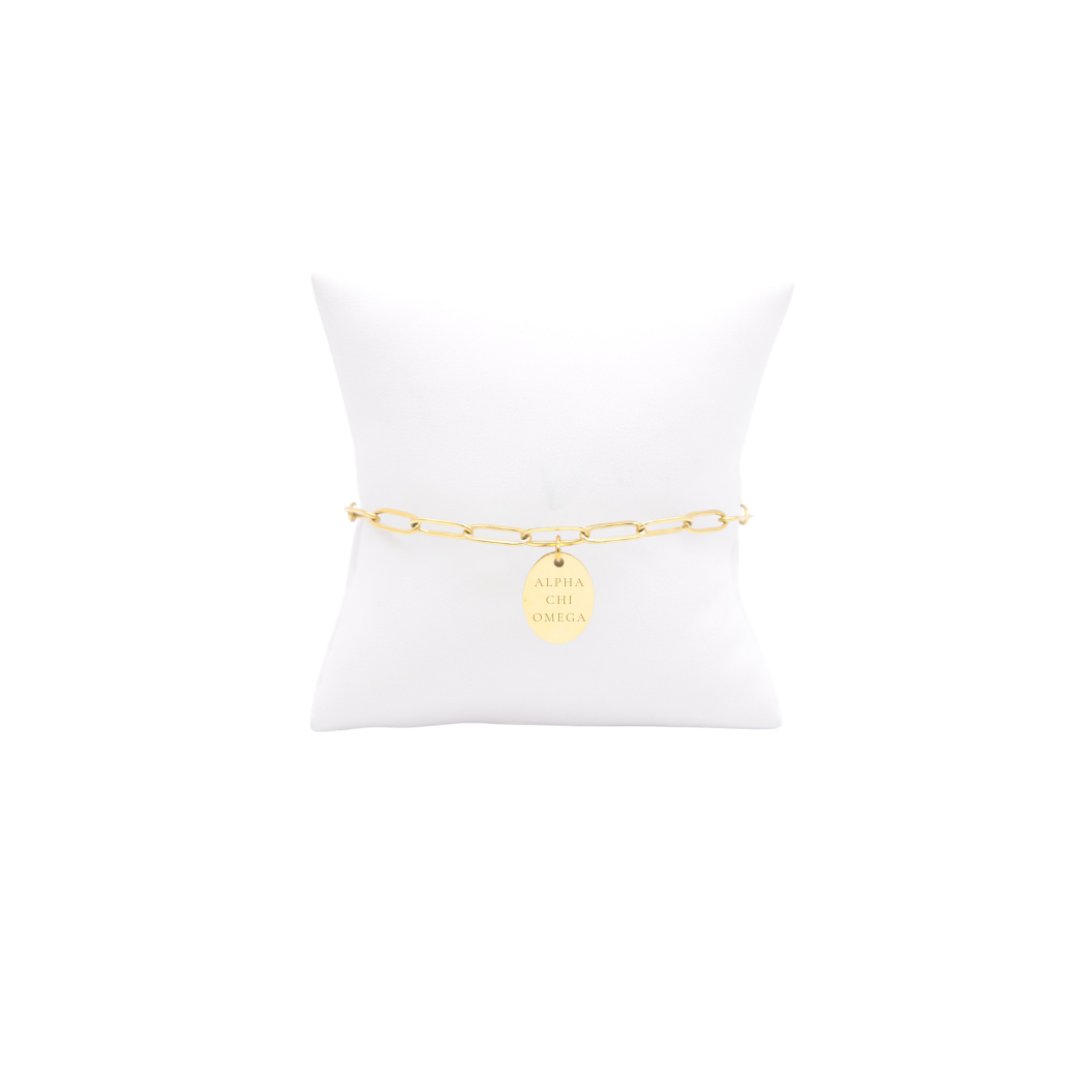 Alpha Chi Omega gold link storyline bracelet with small oval charm with full sorority name on white pillow front view.