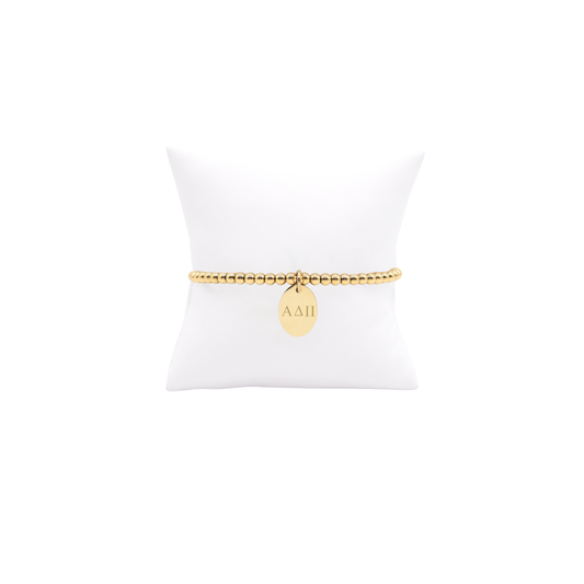 Alpha Delta Pi gold beaded bracelet with small oval charm with horizontal greek letters displayed on white pillow front view.