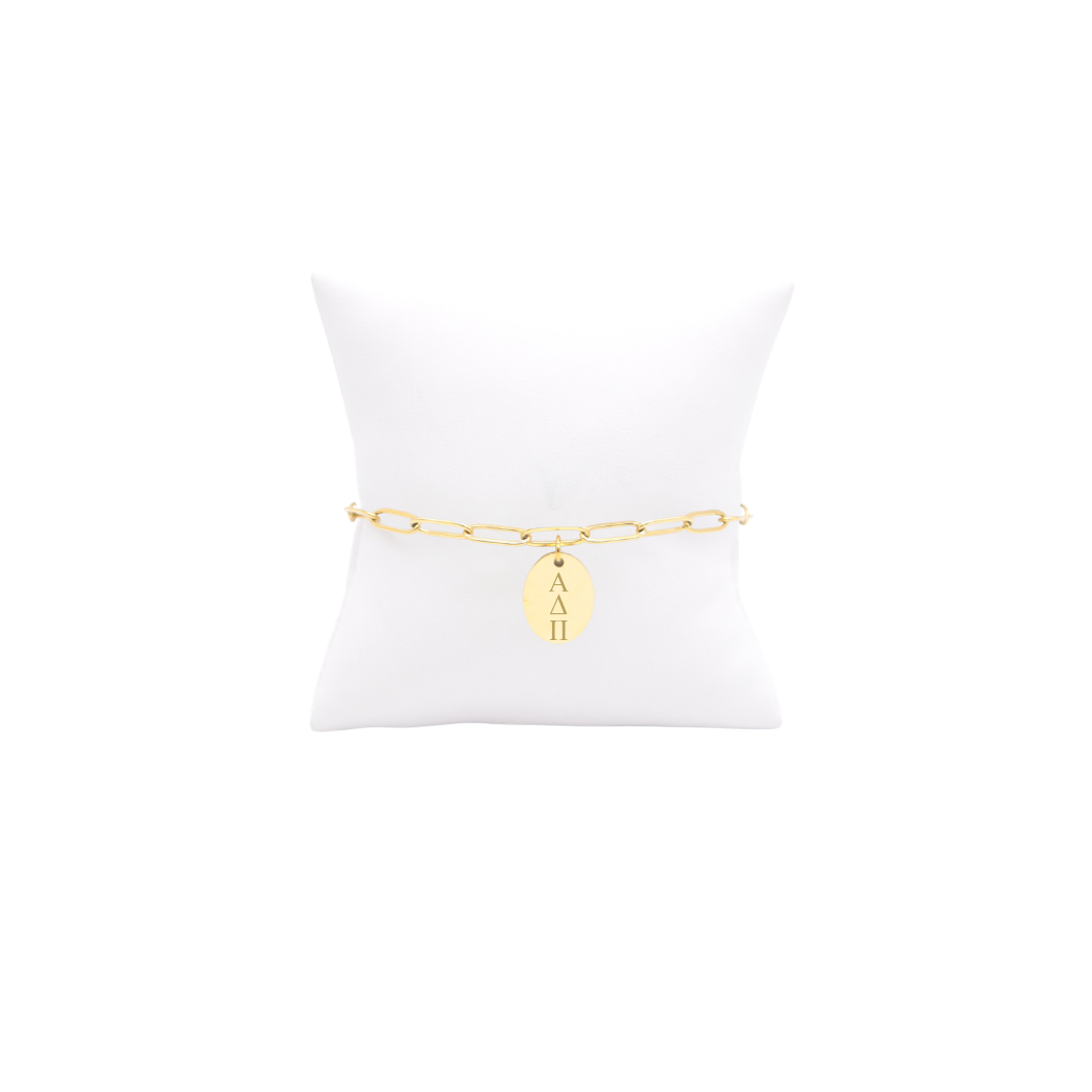 Alpha Delta Pi gold link storyline bracelet with small oval charm with vertical greek letters on white pillow front view.