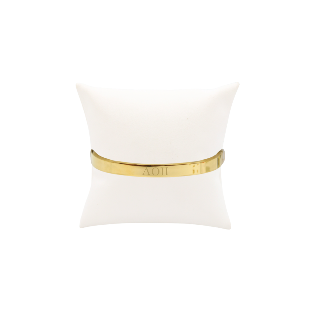 Alpha Omicron Pi gold bangle bracelet with horizontal greek letters on white pillow front view
