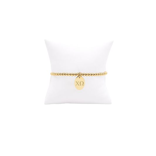 Chi Omega gold beaded bracelet with small oval charm with horizontal greek letters displayed on white pillow front view