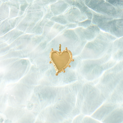 Gold heart-shaped charm featuring a smooth polished center with an organic textured border and decorative bead accents, displayed on a shimmering water background.