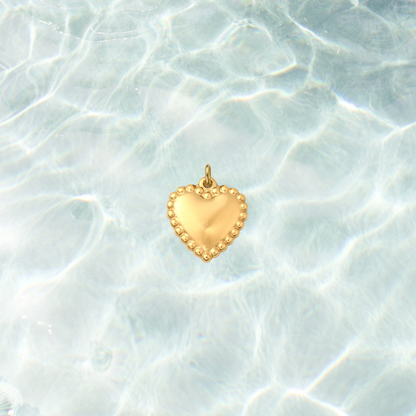 Gold heart charm with a polished center and beaded border on a shimmering water background.