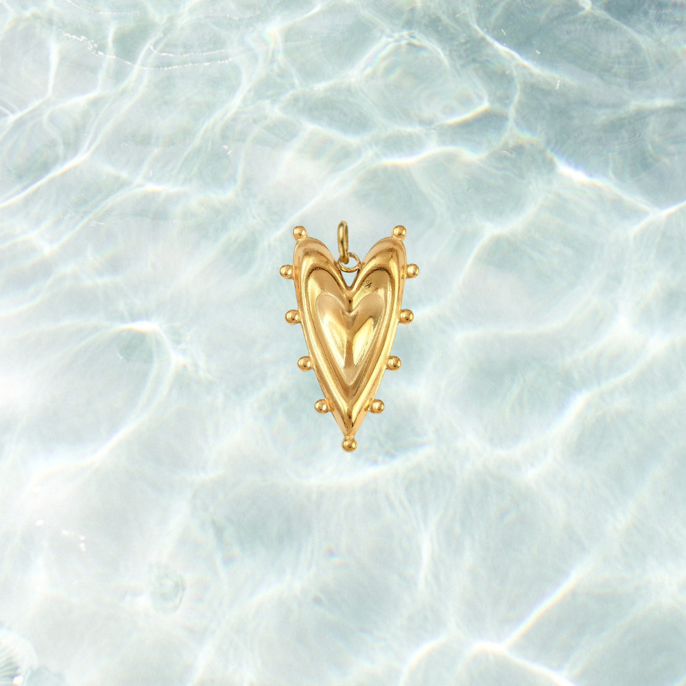 Gold bold heart charm featuring a sculpted puffed heart design with decorative beaded detailing on a shimmering water background.