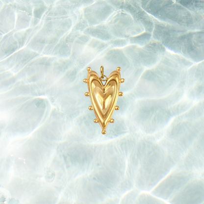 Gold bold heart charm featuring a sculpted puffed heart design with decorative beaded detailing on a shimmering water background.