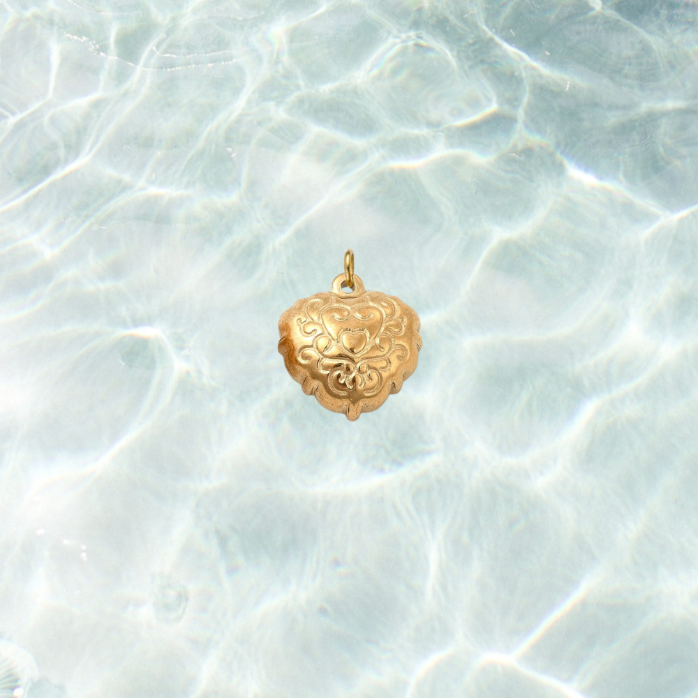 Gold filigree heart charm with an ornate engraved scrollwork design on a shimmering water background.