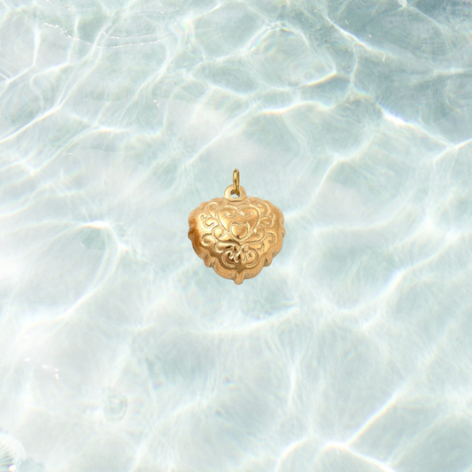 Gold filigree heart charm with an ornate engraved scrollwork design on a shimmering water background.