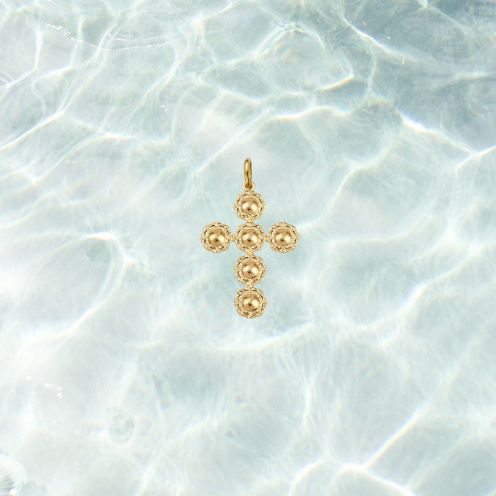 Gold filigree cross charm featuring a textured beaded cross design on a shimmering water background.