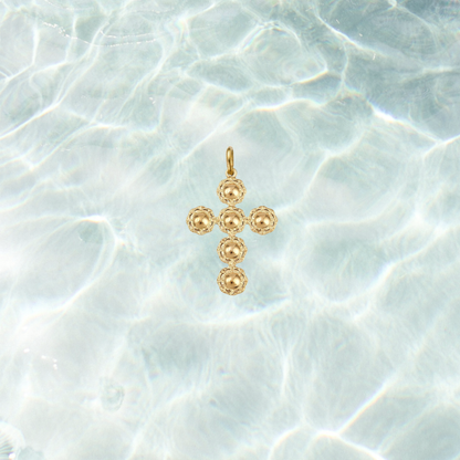 Gold filigree cross charm featuring a textured beaded cross design on a shimmering water background.