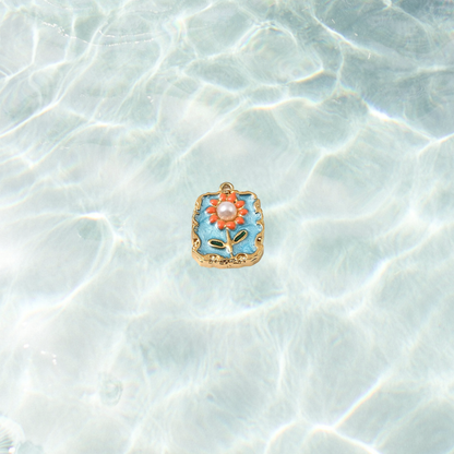 Gold square floral tile charm with pearl tile charm with blue enamel background, orange flower details and pearl center on a waterproof background