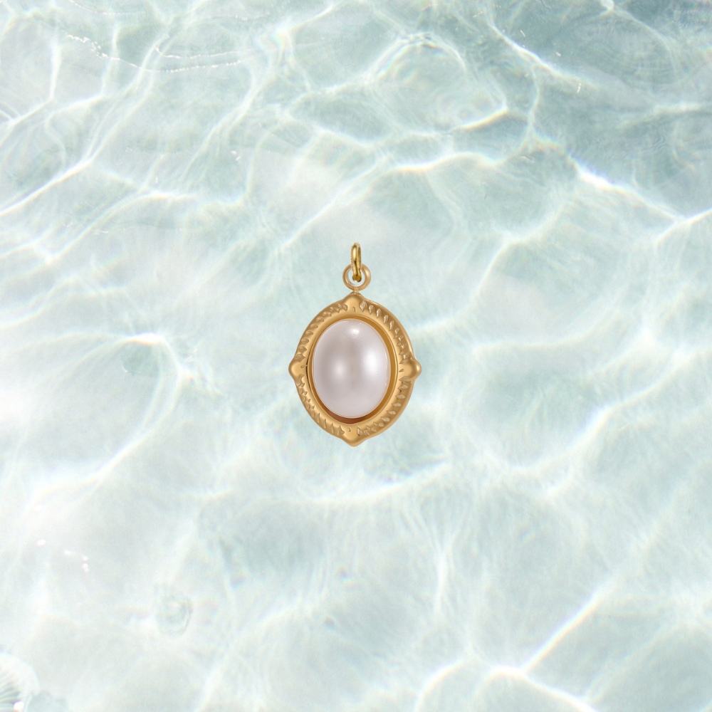Gold oval charm featuring a luminous white pearl cabochon in a decorative bezel setting on a shimmering water background.