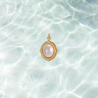 Gold oval charm featuring a luminous white pearl cabochon in a decorative bezel setting on a shimmering water background.