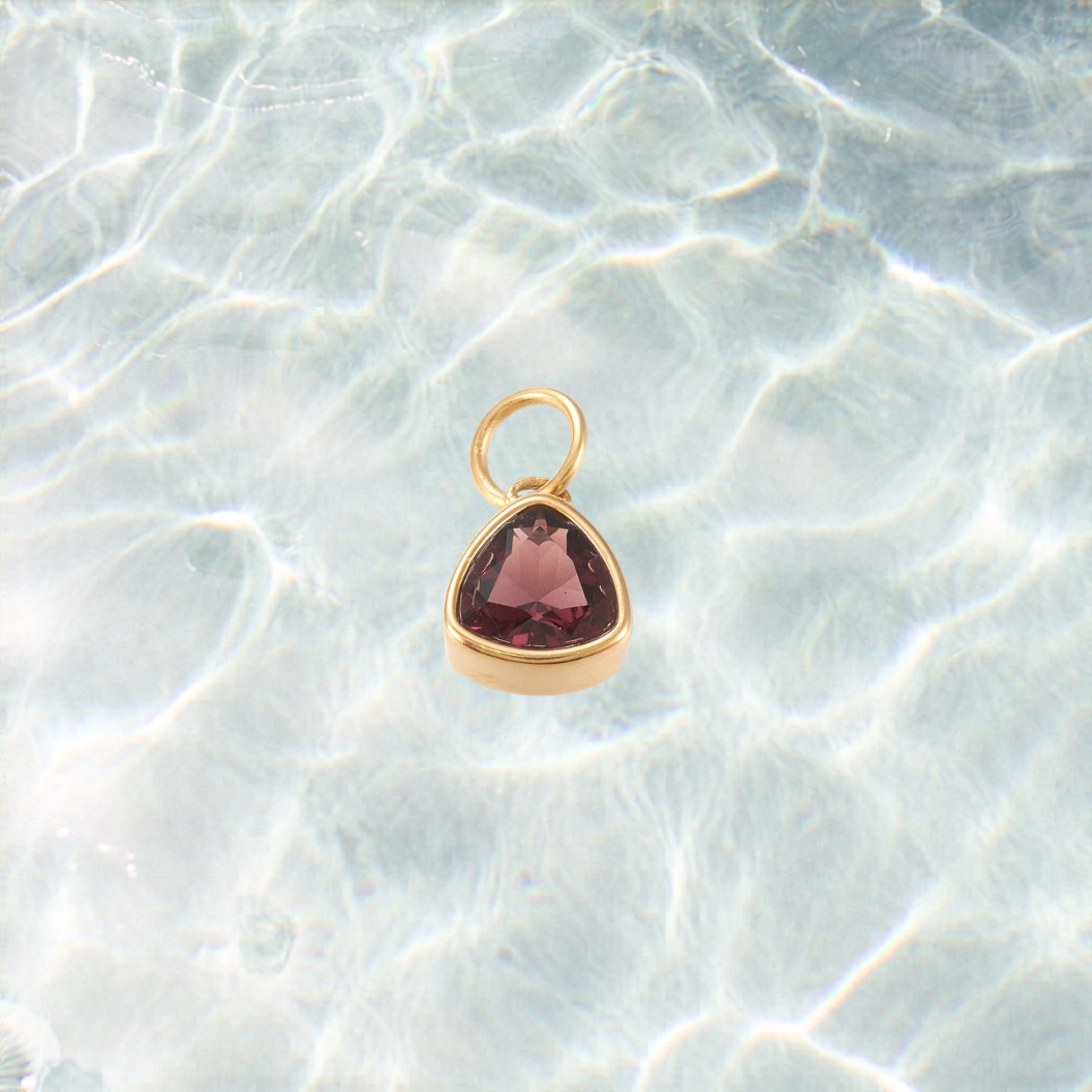 Gold triangular charm featuring a faceted deep burgundy gemstone on a shimmering water background.