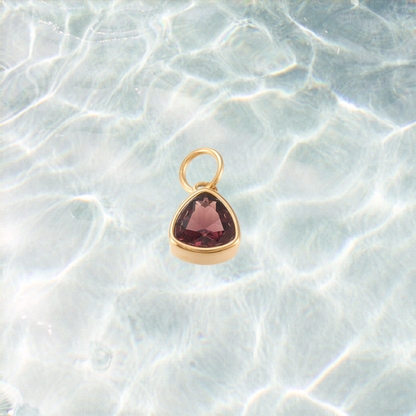 Gold triangular charm featuring a faceted deep burgundy gemstone on a shimmering water background.