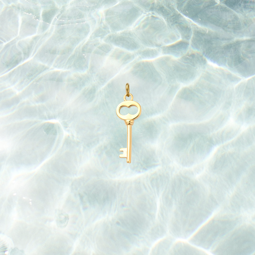 Gold skeleton key charm featuring a heart-shaped bow and polished finish on a shimmering water background.