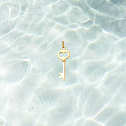 Gold skeleton key charm featuring a heart-shaped bow and polished finish on a shimmering water background.