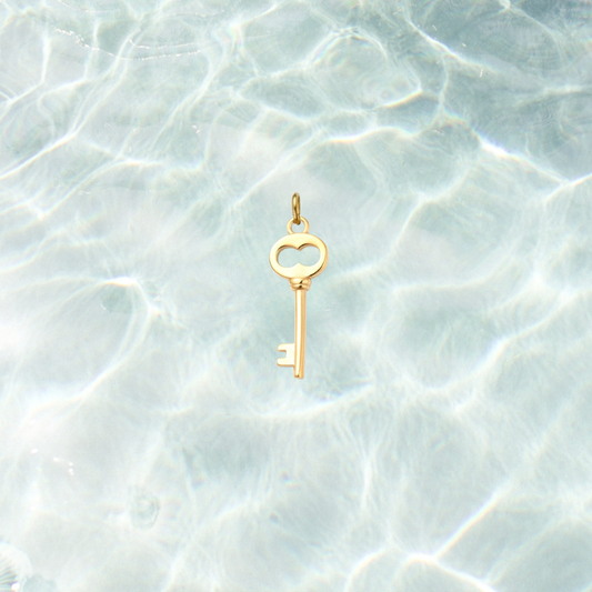 Gold skeleton key charm featuring a heart-shaped bow and polished finish on a shimmering water background.