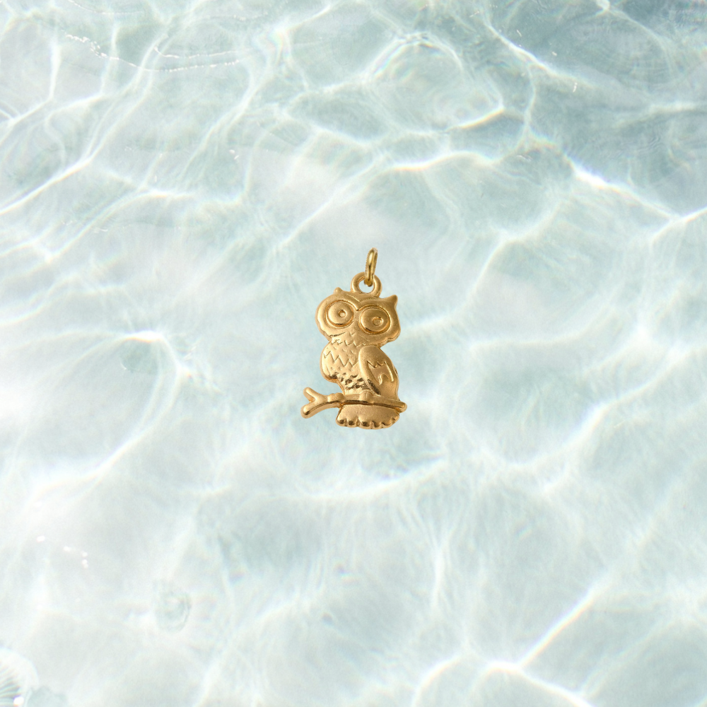 Gold owl charm with engraved feather details perched on a branch against a shimmering water background.