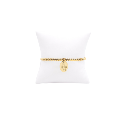Pi Beta Phi gold beaded bracelet with small oval charm displayed on white pillow front view with full new stacked logo
