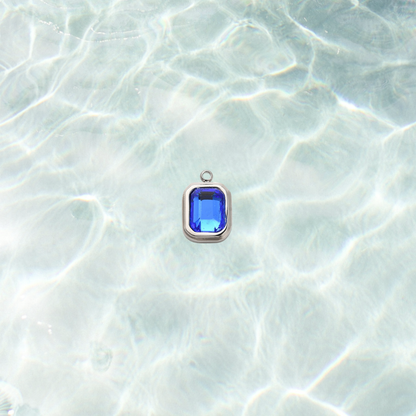 Silver rectangular charm featuring a faceted navy blue gemstone on a shimmering water background.