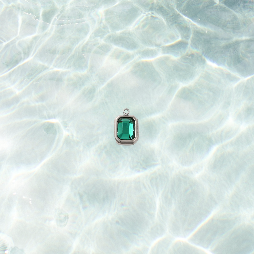 Silver rectangle charm featuring a faceted teal gemstone on a shimmering water bacground.