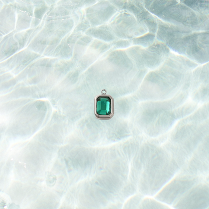 Silver rectangle charm featuring a faceted teal gemstone on a shimmering water bacground.