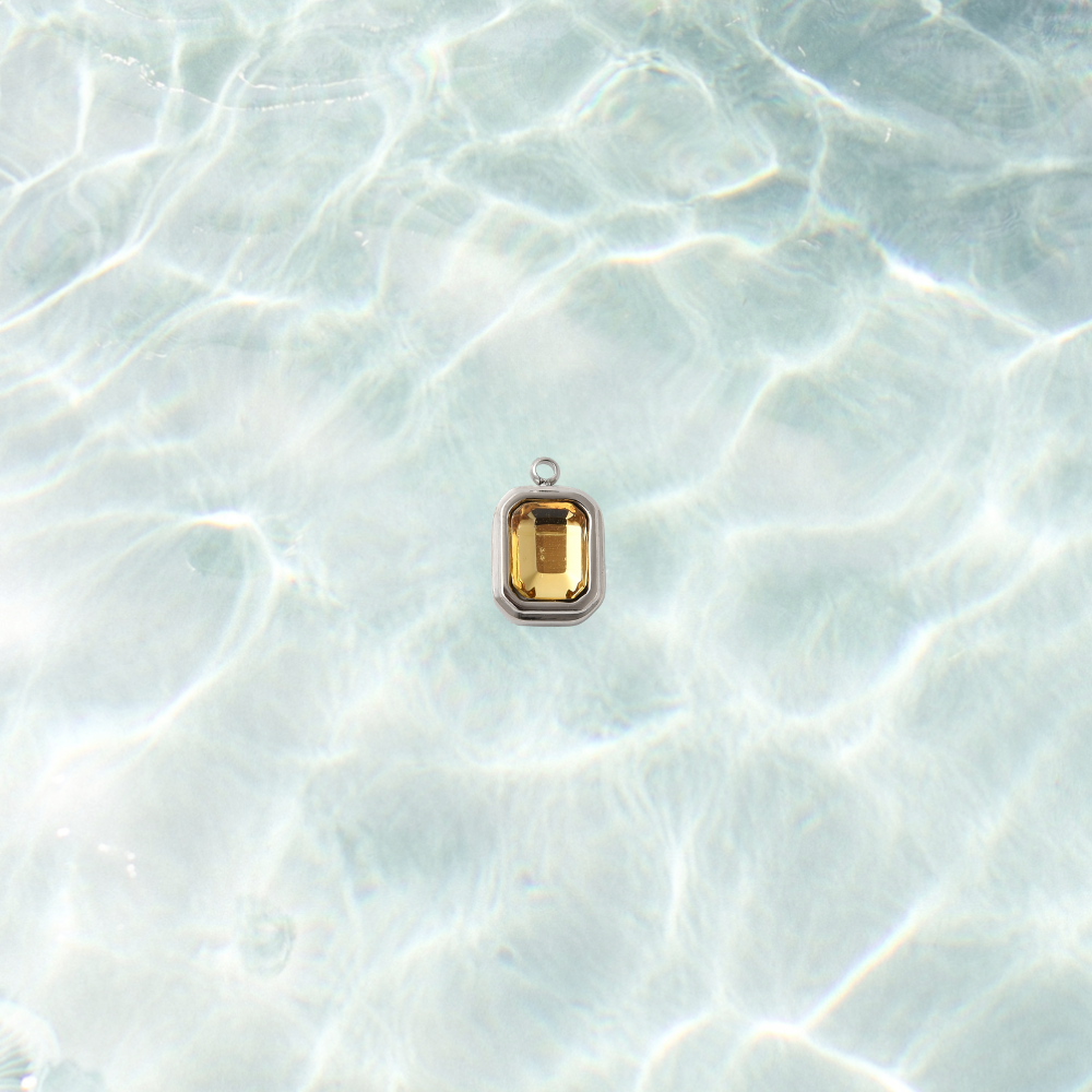 Silver rectangular charm featuring a faceted topaz gemstone on a shimmering water background.