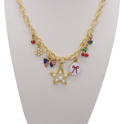 Gold charm necklace featuring a pearl star charm, patriotic bow charm, cherries, pearl cluster, bow illustration charm, and colorful crystal accents displayed on a white jewelry bust