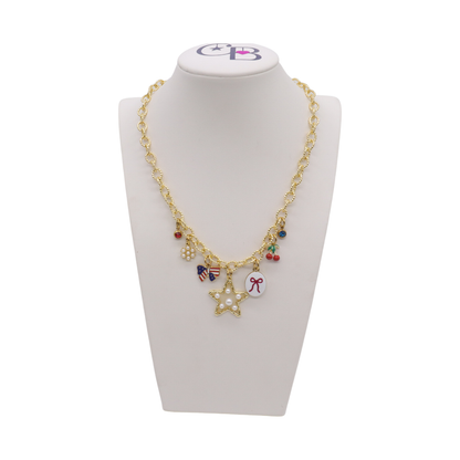 Gold charm necklace featuring a pearl star charm, patriotic bow charm, cherries, pearl cluster, bow illustration charm, and colorful crystal accents displayed on a white jewelry bust