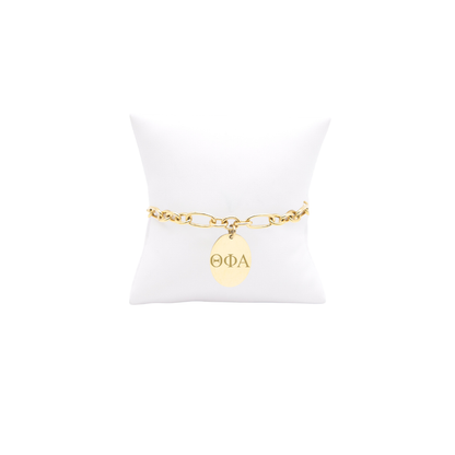 Theta Phi Alpha gold large link keepsake bracelet with large oval charm displayed on white pillow front view with vertical/stacked greek letters